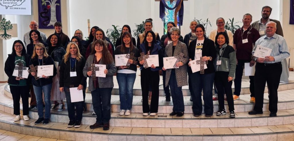 25 educators of various ages stand in front of a church sanctuary holding certificates indicating their years of service at the recent CNH Spring In-Service. Among them are St. Paul Principal Ruth Sebastian (20 years) and Kindergarten Teacher Bethany Bogle (10 years).