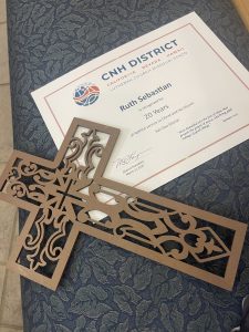 A certificate recognizing Principal Ruth Sebastian for 20 years of service in educator lays flat on chair, stabilized by a wooden filigree decorative cross.