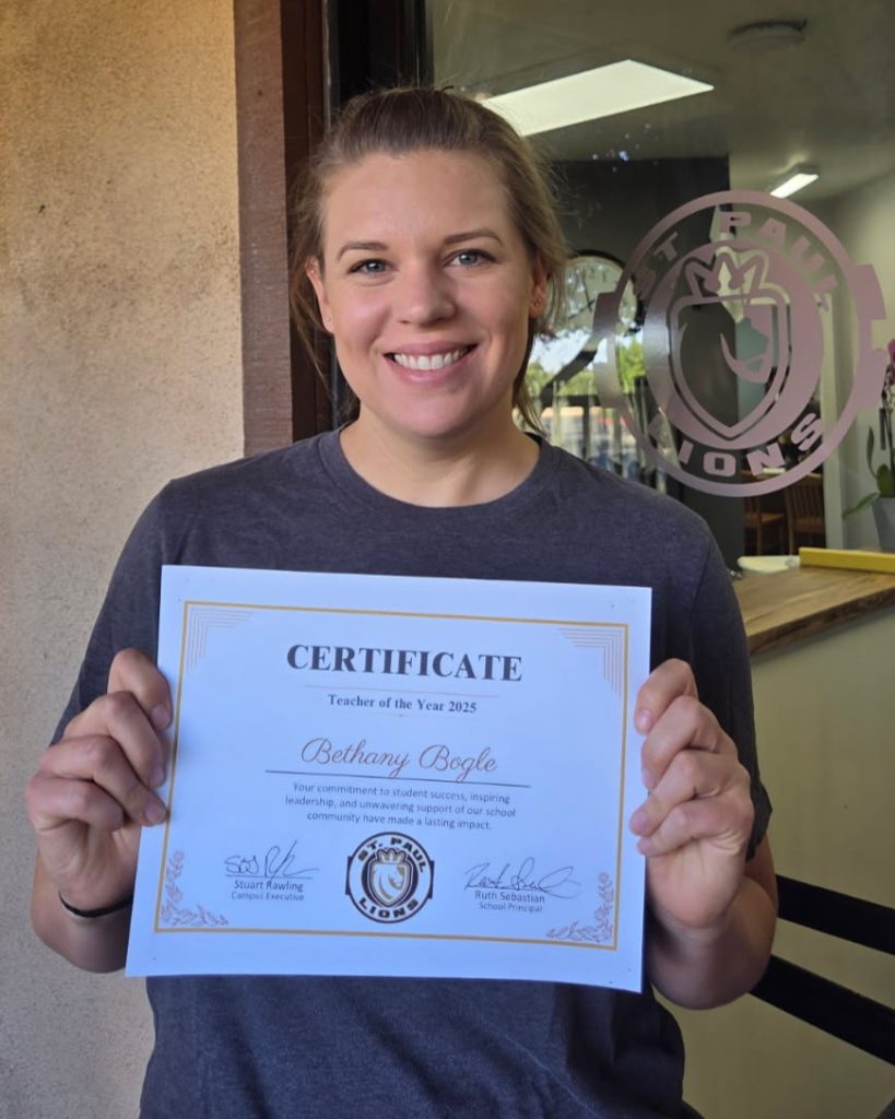 Bethany Bogle smiles while holding the certificate proclaiming her teacher of the year. Mrs. Bogle is wearing her hair pulled back in a ponytail as she stands in front of the office door.