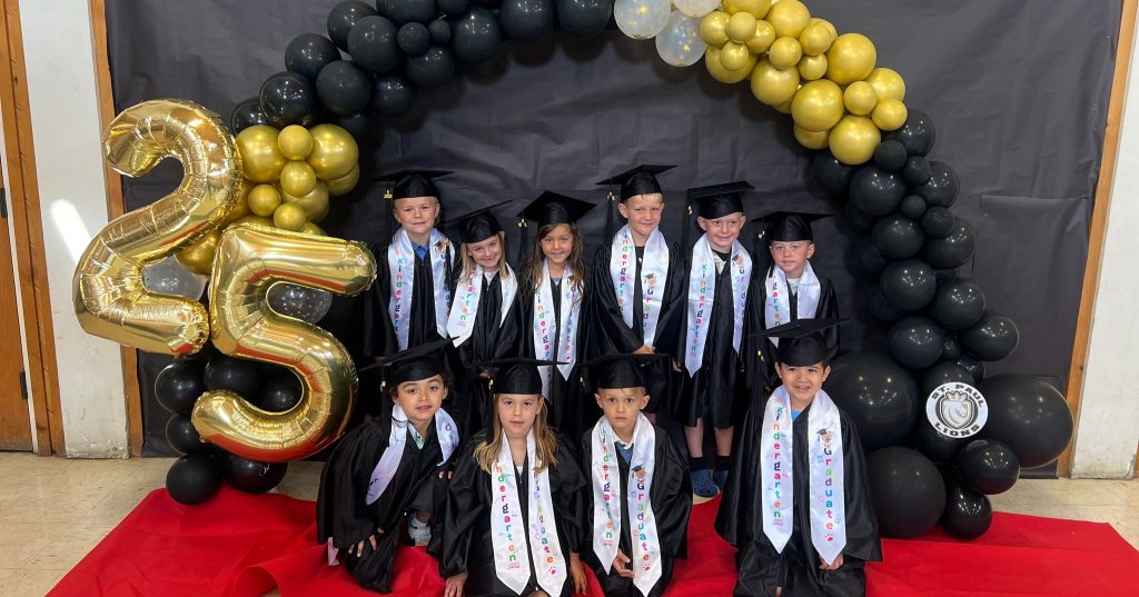 10 kindergartners crouch in front of a black wall decorated with a black, gold and white balloon arch celebrating their promotion to first grade.