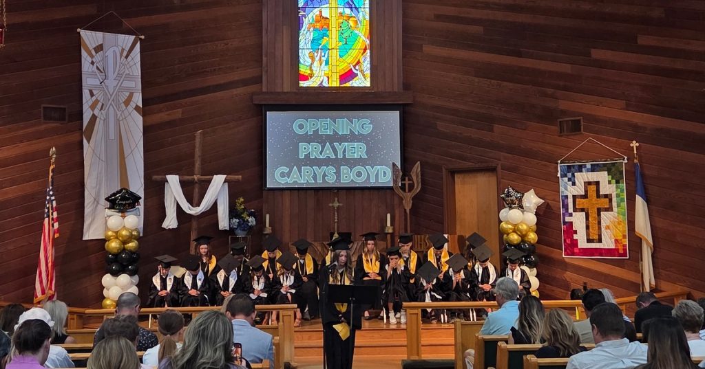Students gather together during a graduation ceremony, with a female student leading peers in prayer.
