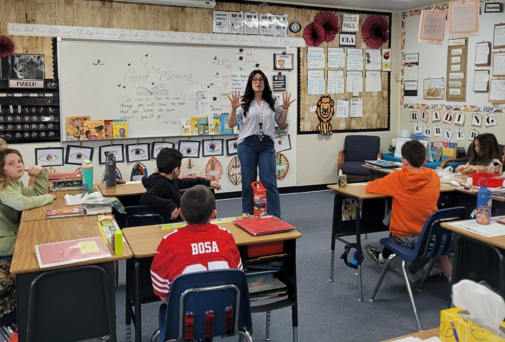 A female teacher stands at the front of a classroom addressing students. She's wearing a white top and blue pants, gesturing with excitement.