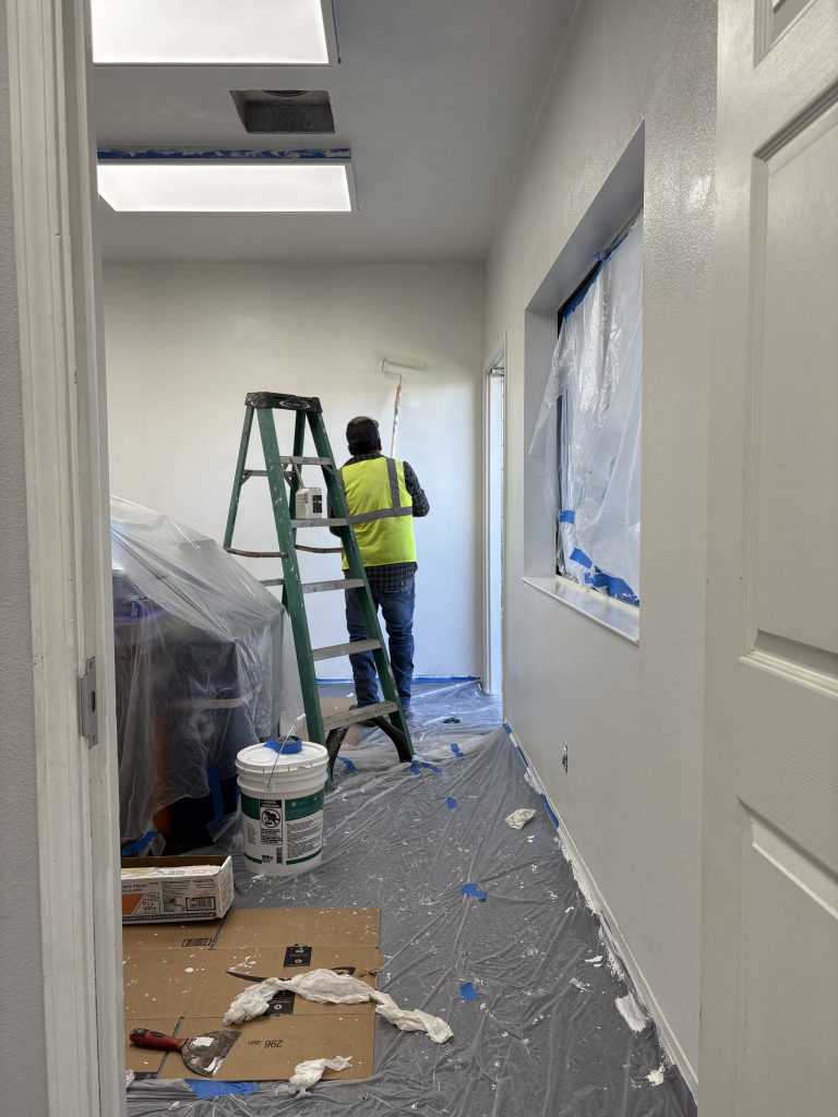 A painter wearing a neon-yellow vest uses a paint roller to paint the teacher's lounge white. A tall ladder is behind him. The rest of the room is covered in plastic sheeting taped down with blue painters tape.