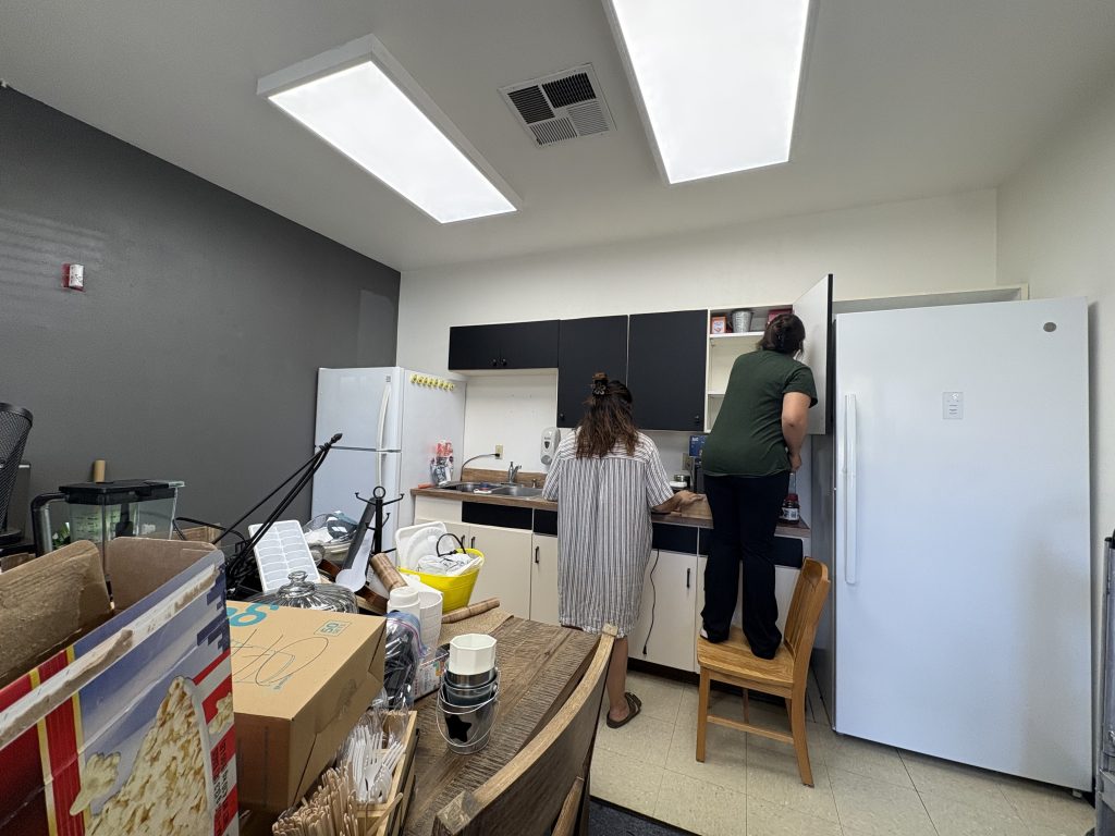 Two women are faced away from the camera. One is standing on a chair as she empties out tall cabinets. The other is standing on the ground beside her to help move items to the table behind them in preparation for the teacher's lounge to be painted.