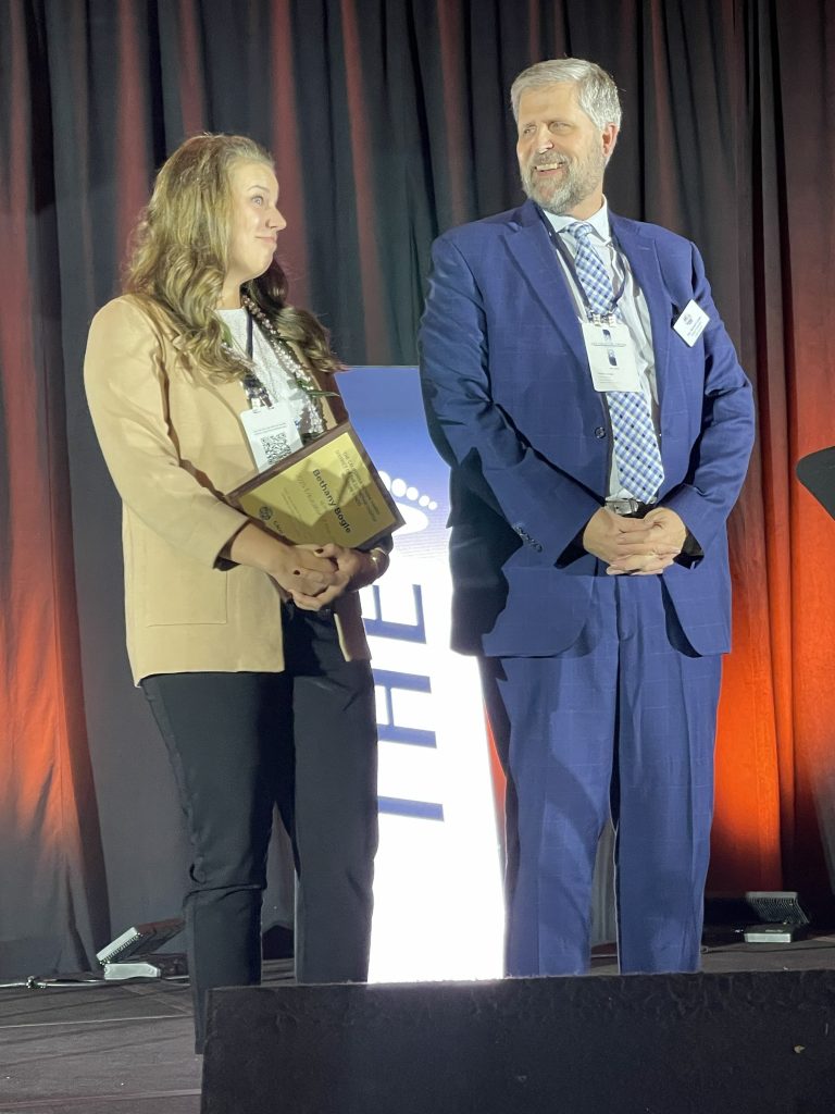 Bethany Bogle, dressed in a beige blazer and black slacks, holds her award plaque next to a CNH district official.