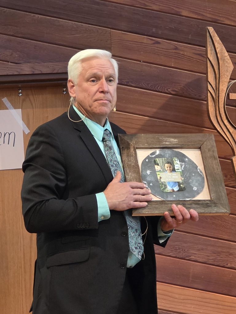 A white-haired man holds a rustic framed image.